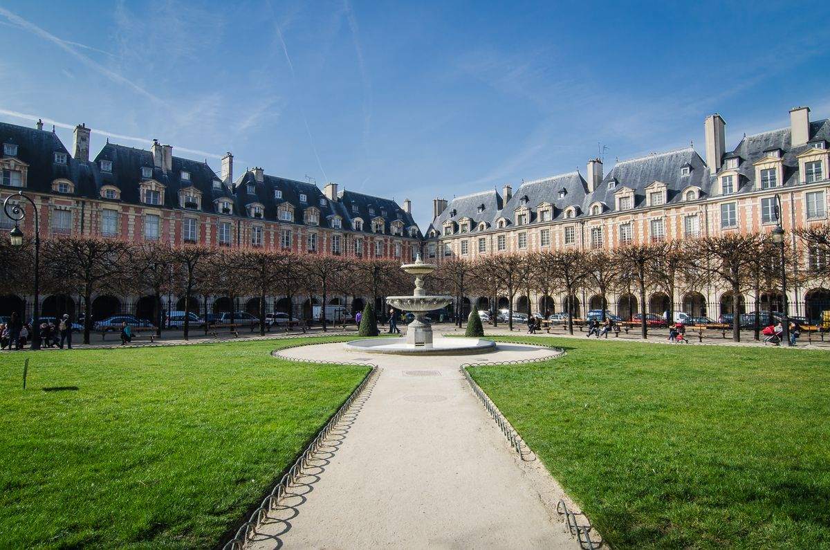 Fontaine au centre d’une place entourée d’arcades, pelouses et bâtiments historiques