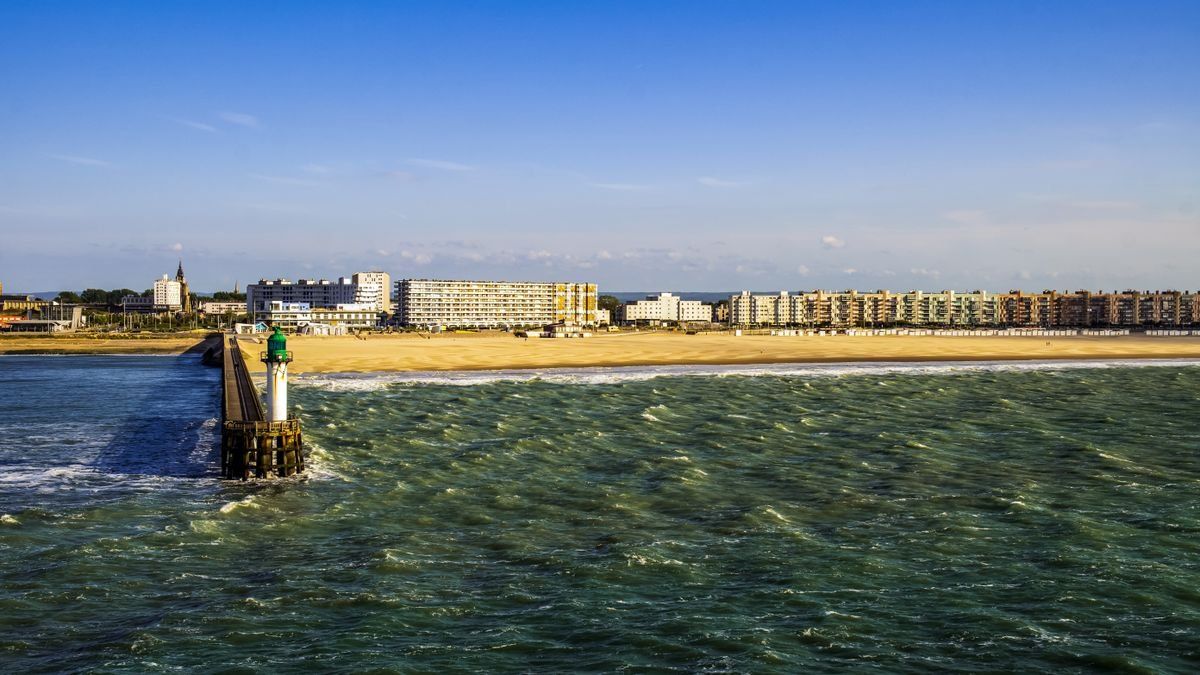 Jetée avec phare vert face à une plage de sable et des immeubles en bord de mer