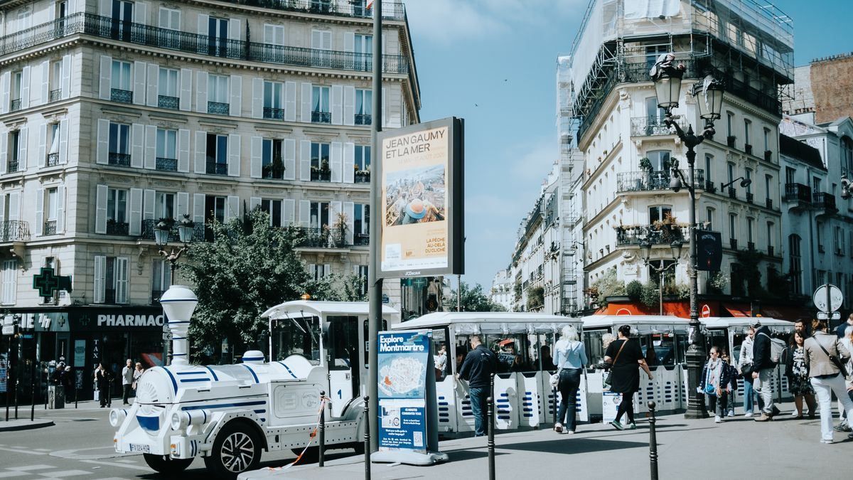 Petit train touristique blanc à un carrefour animé, entouré d’immeubles haussmanniens et de passants